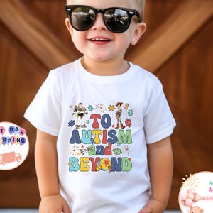 Puede incluir: Camiseta blanca con un gr&aacute;fico colorido con personajes de Toy Story y el texto "To Autism and Beyond".