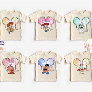 May include: Six beige t-shirts with different Disney characters printed on them. Each character is inside a Mickey Mouse ear silhouette with a watercolour background. The characters are Woody, Jessie, Buzz Lightyear, Mr. Potato Head, and  Rex. The shirts are all the same size and style.