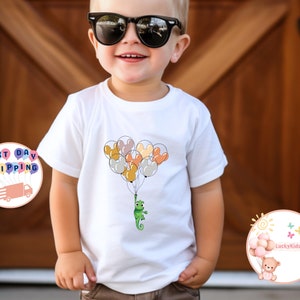 May include: White t-shirt with a green chameleon holding onto a bunch of colorful balloons with Mickey Mouse ears. The balloons are pink, orange, yellow, and gray.
