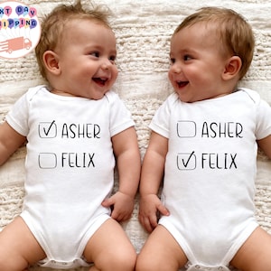 May include: Two smiling babies wearing white onesies. Each onesie has the name "Asher" and "Felix" printed on it, with a checkmark next to one name. The babies are lying on a cream-colored surface.
