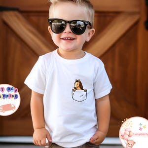 May include: White t-shirt with a pocket design featuring a cartoon horse. The horse is brown and tan, peeking out of a white pocket. The shirt is worn by a young child wearing sunglasses.