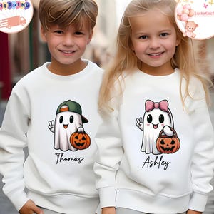 May include: Two white sweatshirts featuring Halloween ghost designs. One ghost wears a green cap and carries a jack-o'-lantern, the other has a pink bow and a jack-o'-lantern. The names "Thomas" and "Ashley" are printed below the designs.