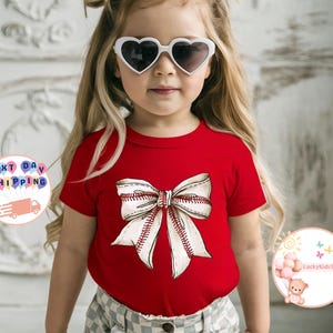 May include: A red short-sleeved t-shirt with a white baseball-stitched bow on the front. The shirt is being worn by a young girl with blonde hair and heart-shaped sunglasses.
