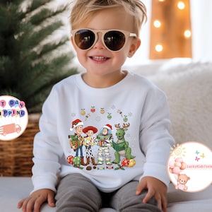 May include: A white sweatshirt with a Christmas-themed Toy Story graphic. The design features Woody, Jessie, Buzz Lightyear, and a green dinosaur, all wearing festive hats. The sweatshirt is worn by a child.