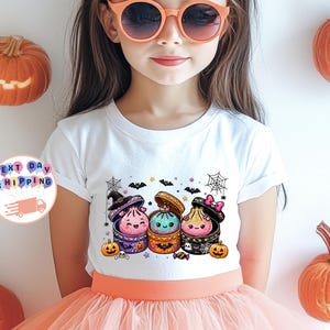 May include: A white short-sleeve t-shirt featuring a graphic of three colorful, glittery dumpling characters inside open round boxes. The design includes small pumpkins, bats, spiderwebs, and stars. The shirt is worn with a peach-colored skirt.