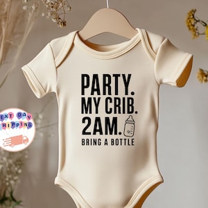 以下が含まれることがあります： クリーム色の半袖ベビー用ボディスーツ。黒い文字で「PARTY. MY CRIB. 2AM. BRING A BOTTLE」とプリントされ、哺乳瓶の小さなグラフィックが添えられています。木製のハンガーに掛けられ、ニュートラルな背景の前に展示されています。