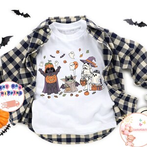 以下が含まれることがあります： ハロウィーンをテーマにしたグラフィックデザインの白いラグランスタイルのシャツ。デザインには、カボチャ姿のダース・ベイダー、ベビーヨーダ、ストームトルーパー、幽霊がすべてコスチュームを着ています。シャツは青と黒のチェック柄のシャツで重ね着されています。