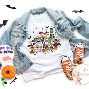 Puede incluir: Camiseta blanca con un colorido dise&ntilde;o de Halloween con personajes de Toy Story. La camiseta se combina con una chaqueta vaquera azul claro y zapatillas deportivas naranjas. Tambi&eacute;n hay murci&eacute;lagos decorativos y una calabaza.