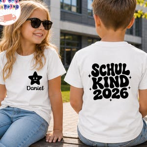 May include: A white t-shirt featuring a black graphic of a smiling star and the name Daniel on the front chest area. The back of the shirt displays the black text SCHUL KIND 2026 in a bold, wavy font, accented by small four-pointed star icons.