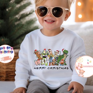 May include: White long-sleeved sweatshirt with a Christmas-themed design featuring the Toy Story characters wearing Santa hats and surrounded by Christmas lights. The text "Merry Christmas" is printed below the characters.