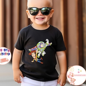 May include: A black t-shirt featuring a colorful graphic of Buzz Lightyear and Woody from Toy Story. The characters are depicted in mid-air. The child is wearing black sunglasses and white shorts.