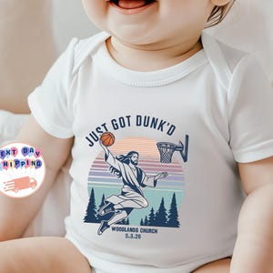 May include: A white short-sleeved baby bodysuit featuring a graphic print of a figure dunking a basketball. The text reads JUST GOT DUNK'D, WOODLANDS CHURCH, and 5.3.26. The design includes a sunset, pine trees, and a basketball hoop in blue and orange.