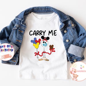 May include: White t-shirt with a cartoon image of a toy character with the text "Carry Me". The character is a spoon with a face, wearing a Mickey Mouse hat and holding a balloon and a chocolate bar.