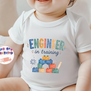 Op de afbeelding: Een wit babyrompertje met korte mouwen en een kleurrijke grafische print. De tekst "ENGINEER in training" staat boven een illustratie van gestapelde blauwe, gele, oranje en groene bouwblokken, een klein grijs tandwiel en een liniaal.