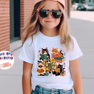 May include: A white short-sleeved t-shirt featuring a graphic print of five colorful cats reading books. The cats are depicted in various poses, some wearing glasses, surrounded by stacks of books, small flowers, and stars. The shirt is paired with blue denim jeans.