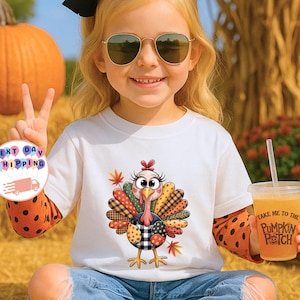 May include: A white short-sleeved t-shirt featuring a graphic of a turkey with colorful patterned feathers in shades of orange, yellow, and black. The turkey has large eyes and is surrounded by autumn leaves. The shirt is worn over an orange long-sleeved shirt.