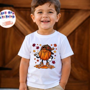 Könnte beinhalten: Ein weißes Kurzarm-T-Shirt mit einem Grafikdruck auf der Vorderseite, der einen baseballförmigen Kürbis mit karierter Kappe und Turnschuhen zeigt, der einen Baseballschläger hält, umgeben von herbstlichen Blättern und orangefarbenen Spritzern.