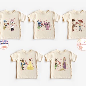 May include: Five cream-colored t-shirts featuring cartoon couples with hearts. Donald Duck and Daisy Duck, Stitch and Angel, Woody and Jessie, Belle and Beast, and Rapunzel and Flynn are depicted. Each design includes red heart accents.