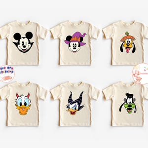 Op de afbeelding: Zes crèmekleurige T-shirts met korte mouwen met cartoonkarakterontwerpen. De ontwerpen omvatten Mickey Mouse, Minnie Mouse met een heksenhoed, Pluto met een pompoenhoed, Donald Duck, een gemene eend en Goofy.