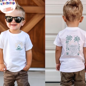 Puede incluir: Camiseta blanca con un diseño de temática playera. La parte delantera presenta un diseño de sol, palmeras y océano. La parte trasera de la camiseta tiene un diseño similar con palmeras, una puesta de sol y un camino sinuoso. La camiseta la lleva un niño pequeño.