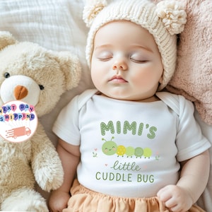 May include: A white short-sleeved baby bodysuit featuring a green caterpillar graphic and the text "MIMI'S little CUDDLE BUG" in green lettering. The garment is worn by a sleeping infant wearing a cream-colored knit hat with two pom-poms.
