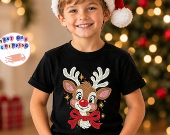 Puede incluir: Una camiseta negra de manga corta con una ilustración gráfica de un reno con nariz roja, astas blancas y pajarita roja, acentuada por pequeñas estrellas amarillas sobre un fondo oscuro.