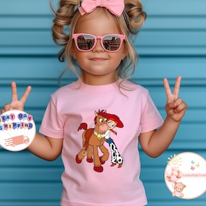 May include: A light pink t-shirt featuring a cartoon graphic of a horse and a cowgirl hugging. The shirt is worn by a young child with pink sunglasses and a pink bow. The background is a blue wall.