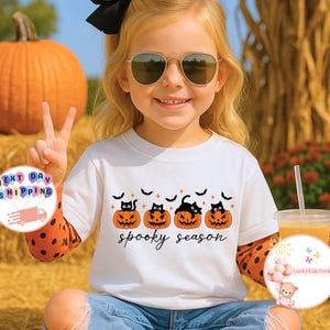 May include: White t-shirt with a Halloween design featuring four jack-o'-lanterns with black cat faces and bats. The words "spooky season" are written in a script font. The shirt is worn by a child.