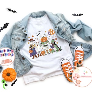 Puede incluir: Camiseta blanca con un diseño de Halloween que presenta personajes disfrazados de Woody, un fantasma, una calabaza, Buzz Lightyear y un dinosaurio. La camiseta se combina con una chaqueta vaquera azul claro y zapatillas naranjas. También hay murciélagos negros recortados y una pequeña calabaza.