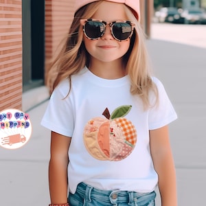 Könnte beinhalten: Ein weißes T-Shirt mit einem Patchwork-Apfel-Design in verschiedenen Mustern und Farben. Das Apfeldesign umfasst florale, gepunktete und karierte Stoffe. Das T-Shirt wird von einer jungen Person getragen, die eine Sonnenbrille und eine rosa Kappe trägt.