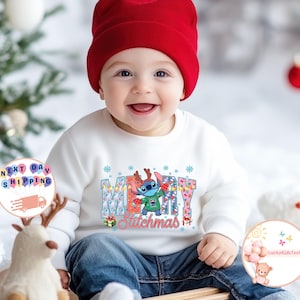 Puede incluir: Un beb&eacute; con una sudadera blanca con un gr&aacute;fico colorido de "Merry Stitchmas". El gr&aacute;fico presenta al personaje Stitch con un su&eacute;ter navide&ntilde;o y cuernos de reno. El beb&eacute; tambi&eacute;n lleva un gorro rojo y vaqueros azules.