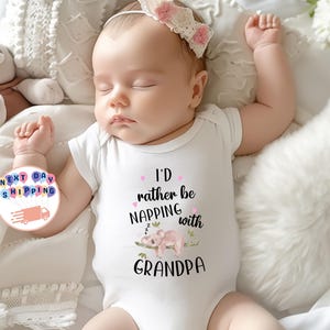 May include: A sleeping baby wearing a white onesie with the text "I'd rather be napping with Grandpa." The onesie features a watercolor illustration of a koala bear. The baby is wearing a white headband with pink flowers.