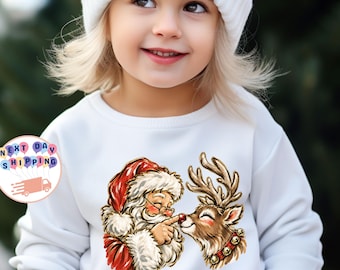 Puede incluir: Una sudadera blanca de manga larga con una ilustración gráfica de Santa Claus tocando la nariz roja de un reno. El reno lleva un collar con cascabeles dorados. La sudadera se combina con un gorro de punto de color blanco.