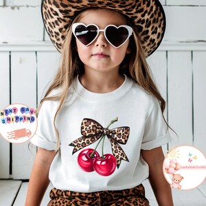 May include: A white t-shirt featuring a graphic of two red cherries with a leopard print bow. The child is wearing heart-shaped sunglasses and a leopard print hat. The shirt is paired with leopard print pants.