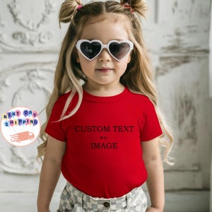 May include: A young child wearing heart-shaped sunglasses and a red t-shirt with the words "CUSTOM TEXT or IMAGE" printed in black. The child is wearing a plaid skirt and has blonde hair styled in pigtails.
