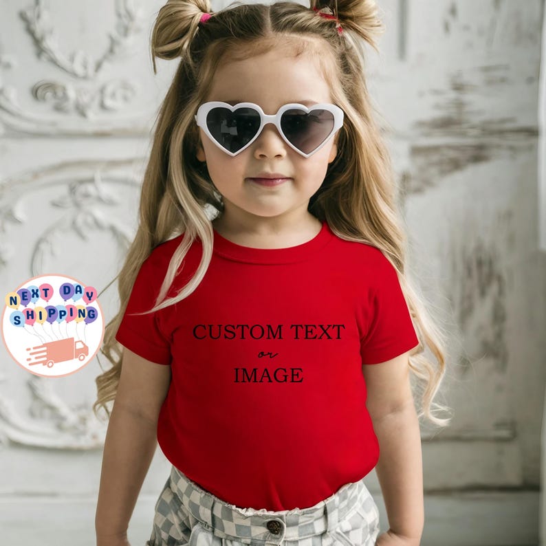 May include: A young child wearing heart-shaped sunglasses and a red t-shirt with the words "CUSTOM TEXT or IMAGE" printed in black. The child is wearing a plaid skirt and has blonde hair styled in pigtails.
