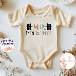 May include: Cream-colored baby bodysuit with short sleeves. The front features the text "MILK, THEN BURPEES" in a playful font, with a barbell graphic above the words. A fun and sporty design for infants.