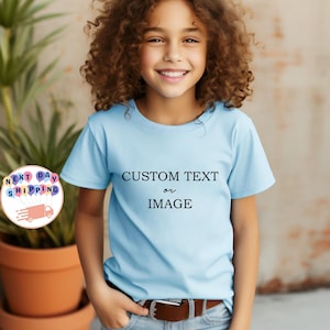 May include: A light blue t-shirt with the text "CUSTOM TEXT or IMAGE" in black. The shirt is worn by a person with curly hair and a brown belt. A logo with balloons and a truck is visible in the background.