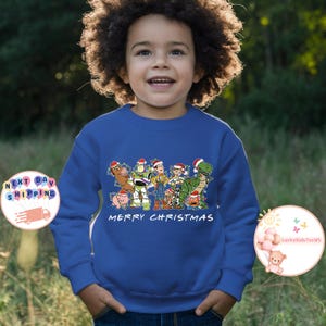 May include: A royal blue sweatshirt with a Christmas-themed design featuring the Toy Story characters Woody, Buzz Lightyear, Jessie, Rex, and Hamm. The characters are depicted wearing Santa hats and are surrounded by Christmas lights. The text "Merry Christmas" is printed below the characters.