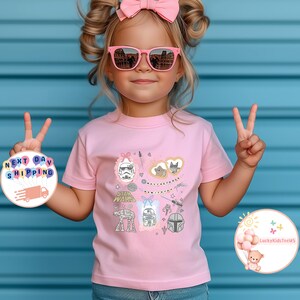 May include: A light pink t-shirt with a Star Wars theme, displaying characters and the phrase "May the Force be with you." The design includes a Stormtrooper, AT-AT, R2-D2, and a Mandalorian helmet. The shirt has a crew neck and short sleeves.