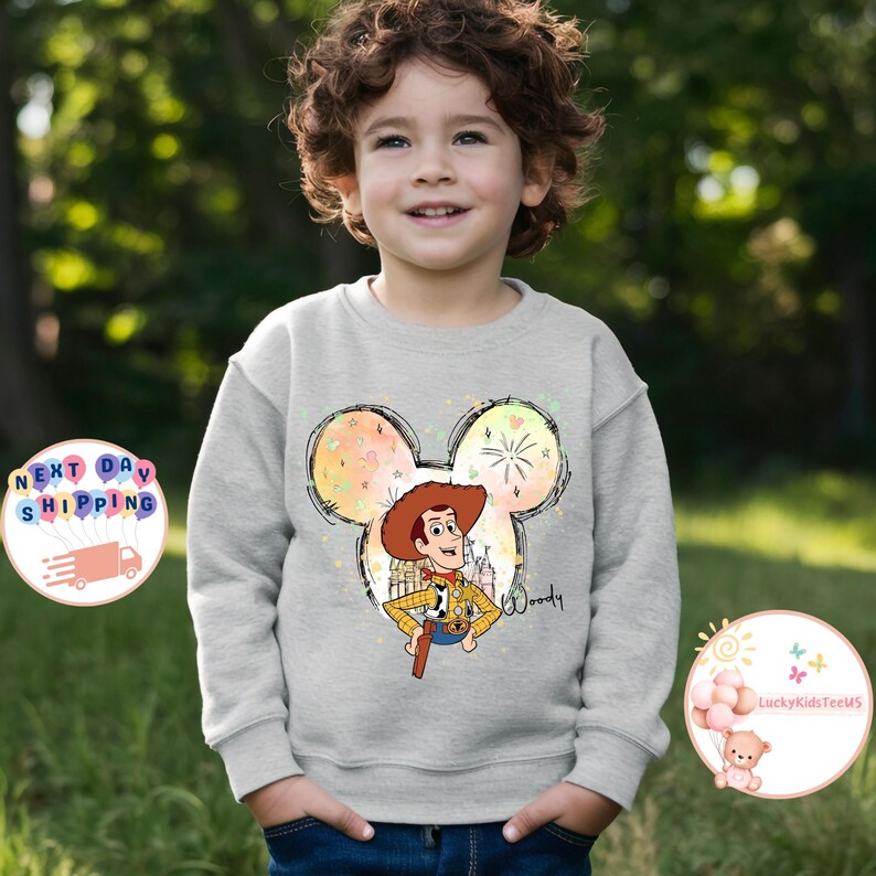 May include: A grey sweatshirt with a colourful Mickey Mouse silhouette featuring a cartoon image of Woody from Toy Story. The text "Woody" is printed on the sweatshirt.