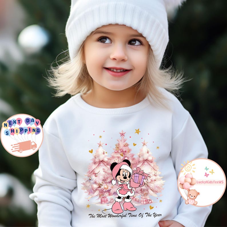 May include: White long-sleeved sweatshirt featuring a Minnie Mouse graphic in a pink Christmas outfit, holding a gift. The design includes pink Christmas trees and the text "The Most Wonderful Time Of The Year". The child is wearing a white beanie.