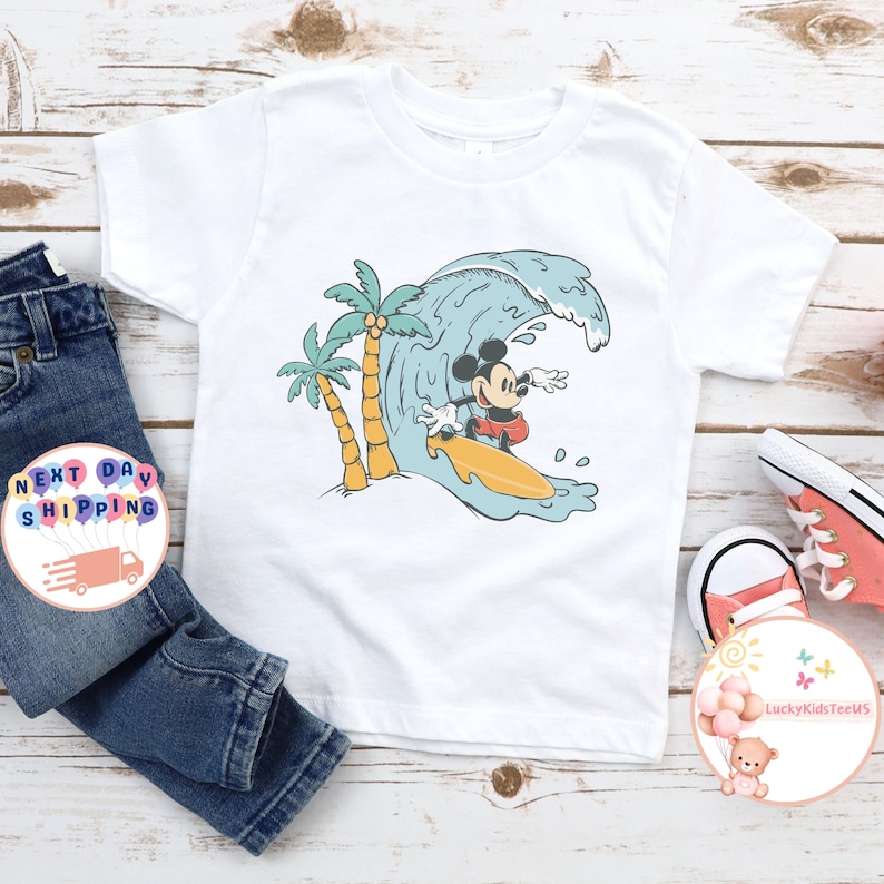 Puede incluir: Camiseta blanca con un gr&aacute;fico de Mickey Mouse surfeando en una ola con palmeras en el fondo.