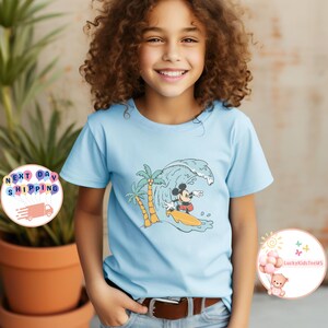 Puede incluir: Una camiseta azul claro con un gr&aacute;fico de Mickey Mouse surfeando en una ola con palmeras en el fondo.