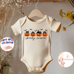 May include: Cream-colored baby onesie with a Halloween design. The design features four orange jack-o'-lanterns with black cat faces and bats, with the words "spooky season" in black script. The onesie is hanging on a wooden hanger.