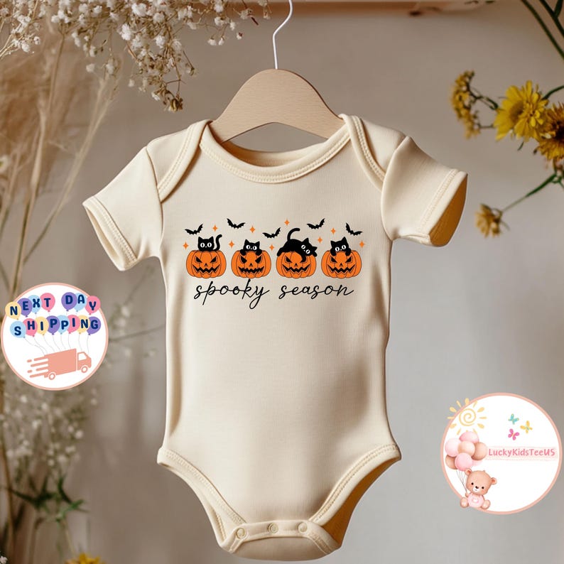 May include: Cream-colored baby onesie with a Halloween design. The design features four orange jack-o'-lanterns with black cat faces and bats, with the words "spooky season" in black script. The onesie is hanging on a wooden hanger.