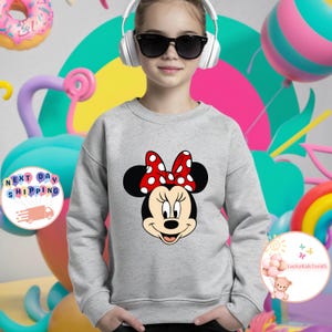 May include: A light grey sweatshirt with a Minnie Mouse graphic. The graphic shows Minnie's face with a red and white polka dot bow. The sweatshirt is a crew neck style. The background includes colourful balloons and a rainbow.