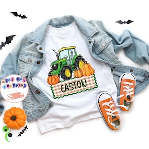 May include: A white t-shirt featuring a green tractor graphic, orange pumpkins, and a brown checkered banner with the name EASTON in green text. The shirt is layered under a light blue denim jacket with orange sneakers and black bat cutouts.
