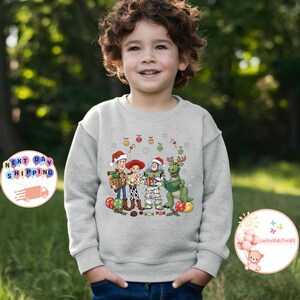 May include: A light grey sweatshirt with a Christmas-themed Toy Story graphic. The design features Woody, Jessie, Buzz Lightyear, and Rex in festive attire, surrounded by baubles and decorations. The sweatshirt is worn by a child.