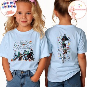 May include: Pale blue Star Wars t-shirt with a festive design featuring characters such as Darth Vader and Yoda. The back of the shirt displays a vertical arrangement of Star Wars figures with Christmas accents. A fun, themed tee for fans.